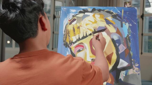 Hind View Of An Asian Artist Boy Holding Paintbrush Mixed Colour And Thinking Before Painting A Girl'S Hair On The Canvas
