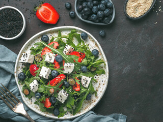 Salad with arugula, feta cheese and berries - strawberry, blueberry, in craft plate on black bacground. Summer salad idea and recipe for healthy vegetarian lunch, dinner. Top view. Copy space.