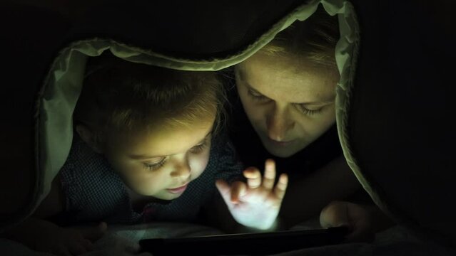 Little girl and her mother using tablet under the blanket. Close-up.