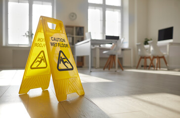 Close up of yellow standing plastic sign with human figure that slips and falls and words Caution Wet Floor placed by janitorial service in empty clean sunny office with desks, chairs and computers