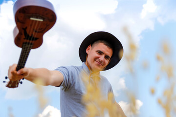 Smiling musician. Young beautiful caucasian man playing ukulele outside. Handsome hipster guy plays ukulele guitar on nature background. Male in a cowboy hat smiles and plays on the guitar