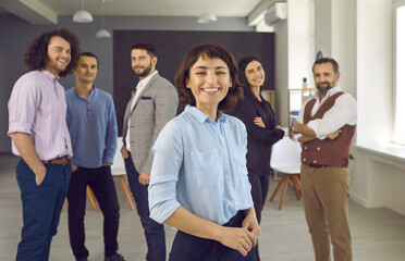 Confident smiling businesswoman standing in office with colleagues in background. Group of successful business people feeling happy and satisfaction. Young female team leader on foreground