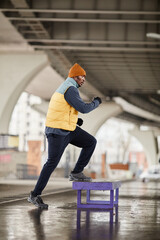 Obraz na płótnie Canvas Side view of young active African American athlete in warm tracksuit, beanie hat and sleeveless jacket exercising on jump box