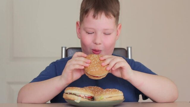 A fat boy eats a hamburger. He is sitting on a chair and very happy.