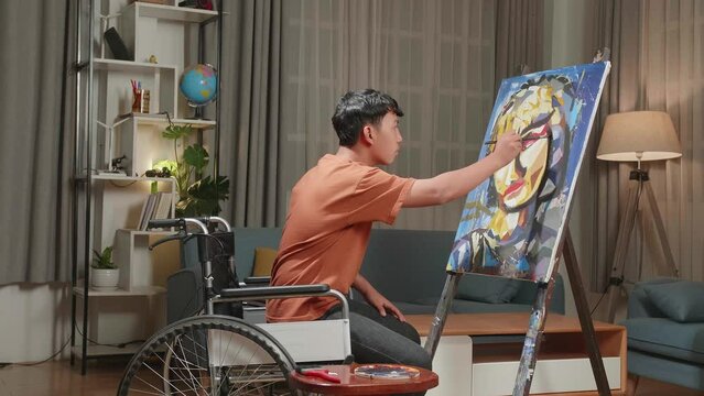 Hind View Side View Of An Asian Artist Boy In Wheelchair Holding Paintbrush Mixed Colour And Painting A Girl On The Canvas
