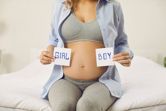 Pregnant woman holding paper cards with inscriptions boy and girl near her bare belly. Cropped image of unknown woman expecting baby sitting on bed and guessing sex of baby. Gender reveal concept.