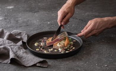 Male hands slice grilled tuna steak with sauce, black round plate. Dark background
