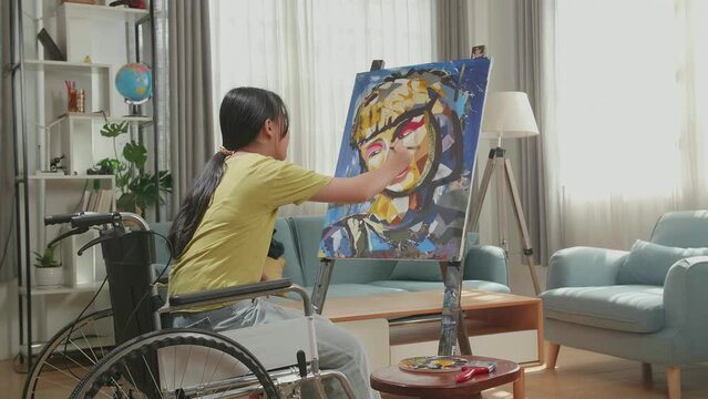 Side View Of An Asian Artist Girl In Wheelchair Holding Paintbrush Mixed Colour Thinking And Painting A Girl On The Canvas
