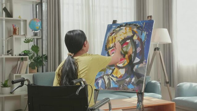 Hind View Of An Asian Artist Girl In Wheelchair Thinking And Painting A Girl'S Face On The Canvas

