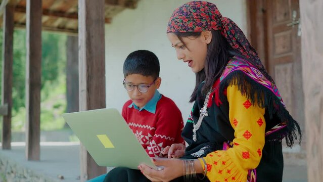 Outdoor shot of a young Indian Asian Rural Woman wearing traditional attire teaching online a little boy sitting together in front of a laptop. concept of Elective home education or Distance Education