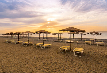  empty beach during beautiful sunrise or sunset with chaise loungues and nice umbrellas with blue sea, sun glow and amazing cloudy sky on thr background