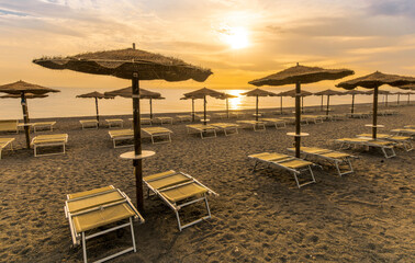  empty beach during beautiful sunrise or sunset with chaise loungues and nice umbrellas with blue sea, sun glow and amazing cloudy sky on thr background