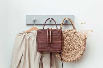 Fototapeta premium A stylish straw bag with a beautiful bouquet and a brown leather backpack hanging on a gray wall next to clothes