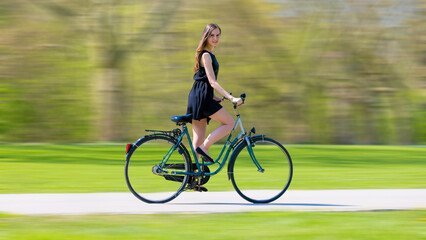 Obraz premium a young woman in a black short dress quickly rides a bicycle on a summer green park