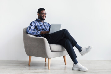 Full length of smiling young black guy with laptop computer sitting in armchair, working online...
