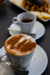 Traditional Italian breakfast. Small white ceramic cups with espresso coffee and cappuccino on a table with saucers and metal spoons at a local bar (European coffee shop) in Milan, Lombardy, Italy. 