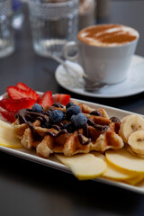 Traditional Italian breakfast. Fresh classic waffles with fruits, sugar powder, chocolate and cappuccino coffee with cinnamon in Milan, Lombardy, Italy. European food, drinks  pastry at a local bar.
