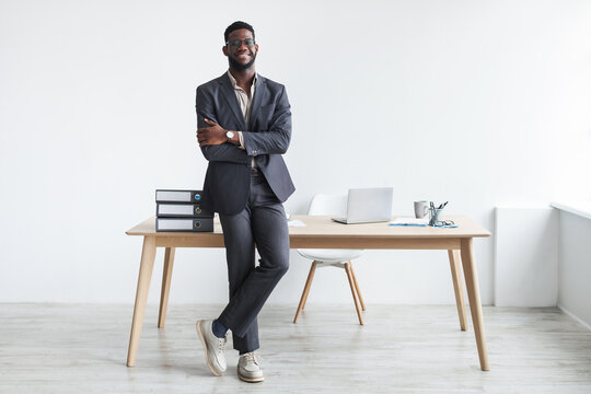 Full length of happy young black businessman in formal suit standing with crossed arms near office desk, copy space