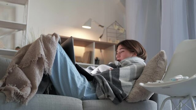 Covid disease. Sick woman. Home recovery. Ill lady wrapped plaid laying sofa looking funny movie on laptop in light room interior.