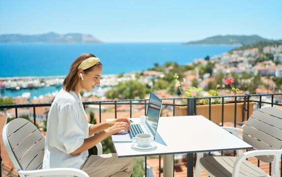 Young woman using laptop computer at cafe balcony of resort hotel with sea view, working typing emails browsing online enjoying drinking coffee
