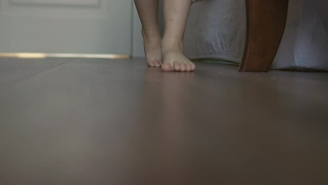two barefoot legs of little child stepping and walking into camera on brown wooden floor at home indoors. close-up view naked feet steps kid toddler. 