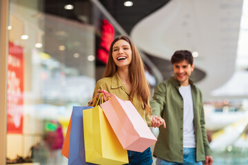 Joyful Wife Carrying Bags Pulling Husband Shopping Together In Mall