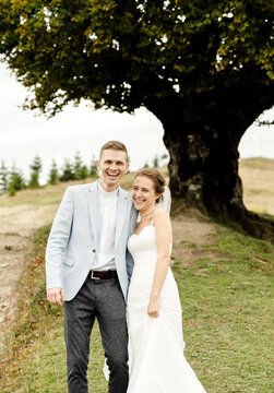 a cheerful wedding couple walks under a large and beautiful branchy tree in the mountains. wedding day