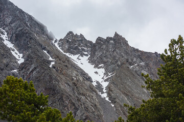 Gloomy mountain landscape with high sharp rockies with snow in gray low clouds. Closeup of sharp rocks and peaked tops in gray cloudy sky. Bleak overcast scenery with mountain range with pointy rocks.