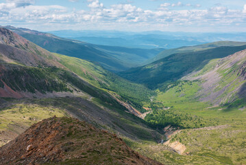 Scenic aerial view from mountain pass to green forest valley among mountain ranges and hills on horizon at changeable weather. Green landscape with sunlit mountains under cumulus clouds in blue sky.