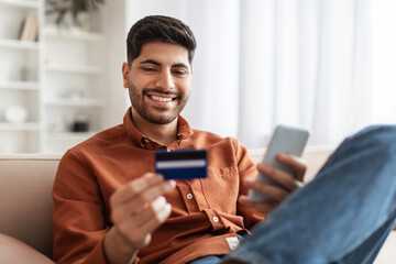 Smiling Arab guy using phone and credit card at home