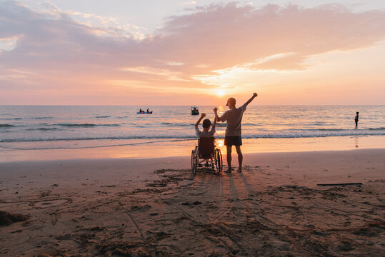 Behind of young man with disability and parent or volunteer or caregiver looking sunset on the sea beach at sunset with travel in summer, Positive photos give life energy and power concept.