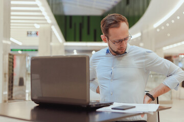 Man holds on to his back, suffers from lower back pain, working on computer. Sedentary work office worker - obrazy, fototapety, plakaty