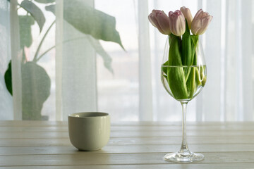 Bouquet of pink tulips in glass vase and cup of coffee on white wooden table near the window