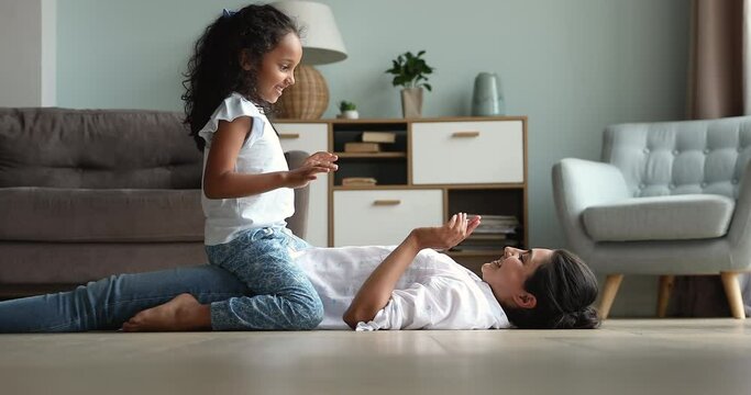 Cute Indian girl play patty cake game with mother, woman lying on warm floor in living room, clapping hands, enjoy free carefree playtime with 6s preschool daughter. Fun, leisure, family bond concept