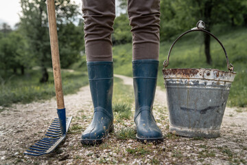 Close up on legs of unknown woman in rubber boots standing on the dirt road with metal bucket and rake beside - farm agriculture and sustainable life concept