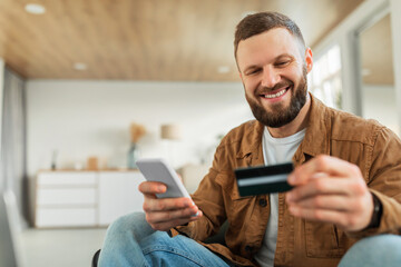Happy Male Shopping Using Smartphone And Credit Card Sitting Indoors - obrazy, fototapety, plakaty