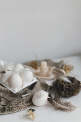 Fototapeta na wymiar Easter rustic still life. Easter natural eggs in tray, feathers, willow branches, nest on burlap on white wooden table. Simple rural Easter aesthetics in pastel beige colors. Happy Easter