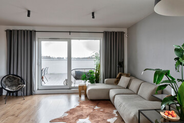 Modern living room in stylish interior with large window. Comfortable sofa and armchair on wooden floor and green plant in the room in luxurious apartment.