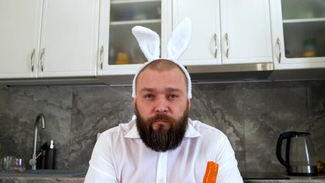 A brutal man with a beard and bunny ears looks into the camera and eats a carrot in the kitchen. Easter bunny, humor