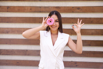 Portrait of a Caucasian girl in white clothing with a flower in front of her eye, showing the okie sign with her hands on a brown ribbed background with copy space