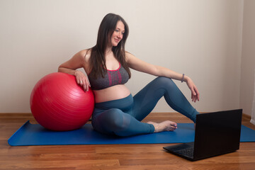 Fototapeta na wymiar Fitness pregnant woman doing yoga workout on yoga mat opposite laptop with online master class. Healthy sports pregnancy concept. 