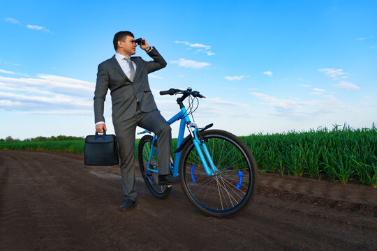 business concept - A businessman looking through binoculars into the distance, poses with a bicycle on a green grass field, dressed in a business suit, he has a briefcase and documents