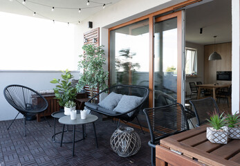 Beautiful terrace with rattan and wooden furniture, chair, table and sofa with pillow. Large window and lamps outdoor. Modern patio with plants, stylish design for relax end rest time.