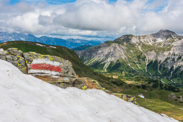 Red and white marking of the hiking trail in the Austrian Alps. Sunny mountain panorama with snow in the foreground. - 485427523