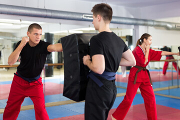 Caucasian man exercising jabs with teenage boy who holding punching pad during group karate...