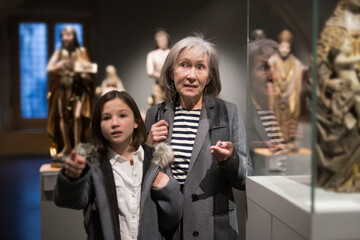 Friendly elderly female tutor showing interested tween girl exposition of artworks in museum of...