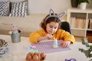 Happy little girl with bunny ears painting the clay rabbit preparing for Happy Easter day. Preparing handmade Easter decoration at home.