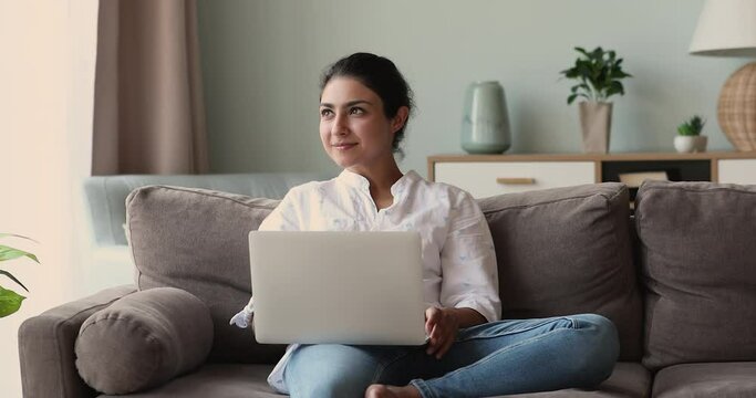Young Indian freelancer sit on sofa with laptop working at home. Beautiful woman put wireless computer on laps smile typing text, do telework, chatting on-line, share messages in social media concept