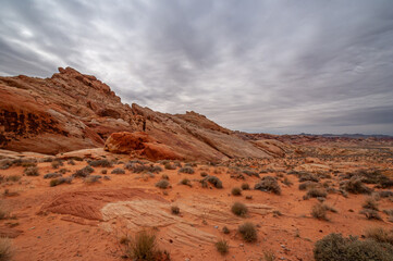 Fototapeta na wymiar Overton, Nevada, USA - February 25, 2010: Valley of Fire. Wide landscape with slanted mountain outcrop raising out of dry red rock desert floor with shrubs under heavy gray cloudscape.