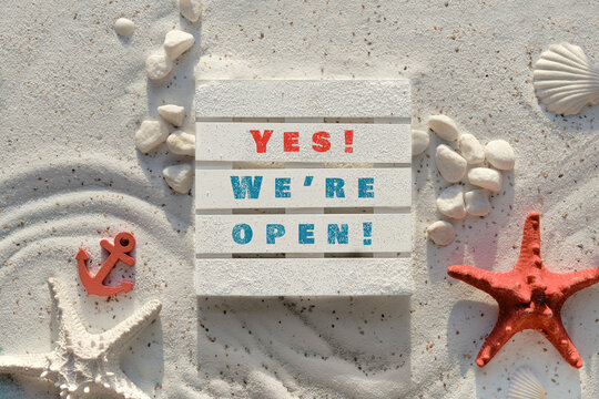 Sign yes we are open on wooden palette. Sand background with pebbles, stones, red starfish and anchor. Sand and sea decor background.